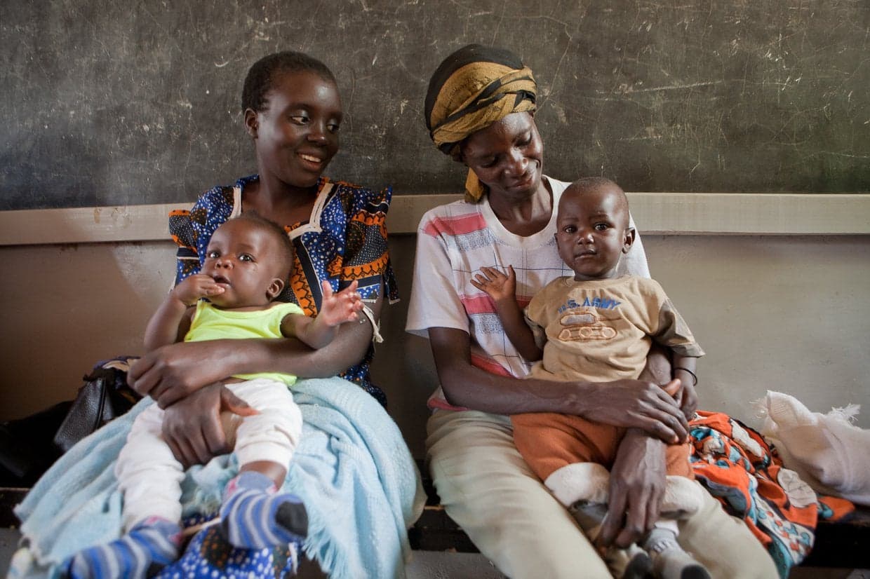 At a clinic in Kenya, two women sit on a bench holding babies on their laps.
