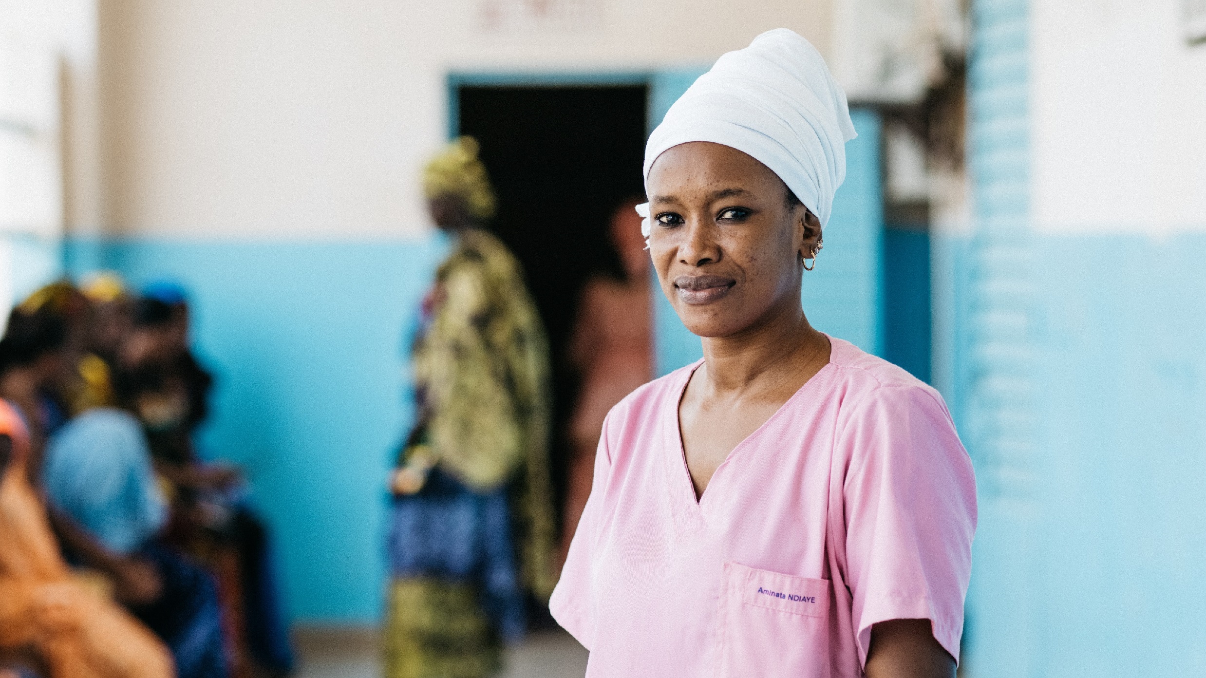 A health worker wearing pink scrubs and a white headwrap looks at the camera.